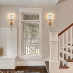 Clérachic pair of brass-gold wall sconces with glass shades either side of a tall white-framed window in a hallway.