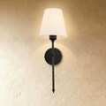 Clérachic black wall sconce with a white tapered fabric shade on a slim rod and round backplate, glowing on a limewash wall.
