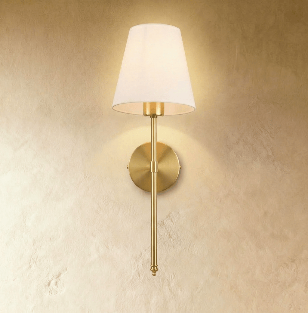 Clérachic brushed gold wall sconce with a white tapered fabric shade on a long slim rod, lit warm on a plaster wall.