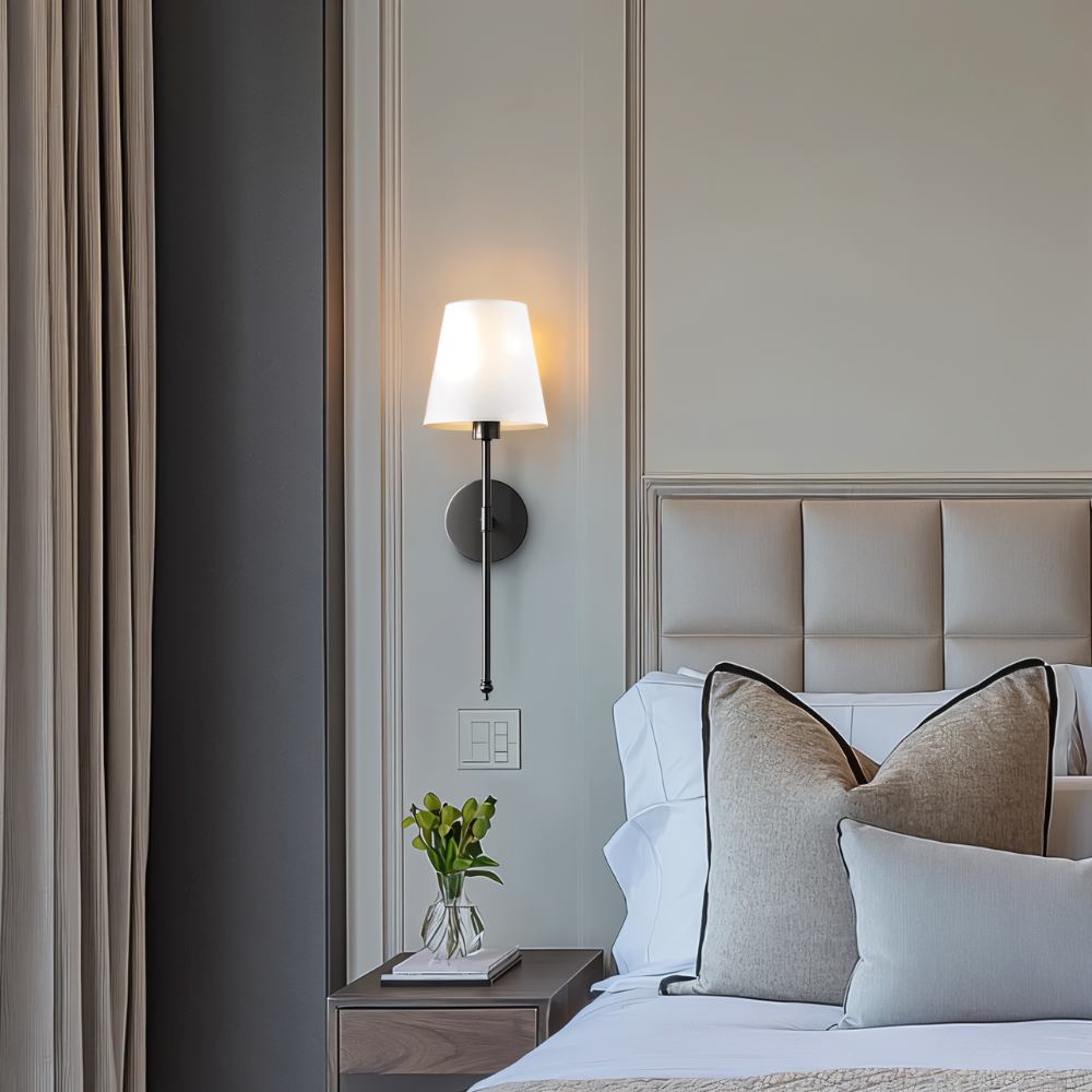 Clérachic black wall sconce with a white tapered shade beside a panelled headboard wall, above a bedside table.