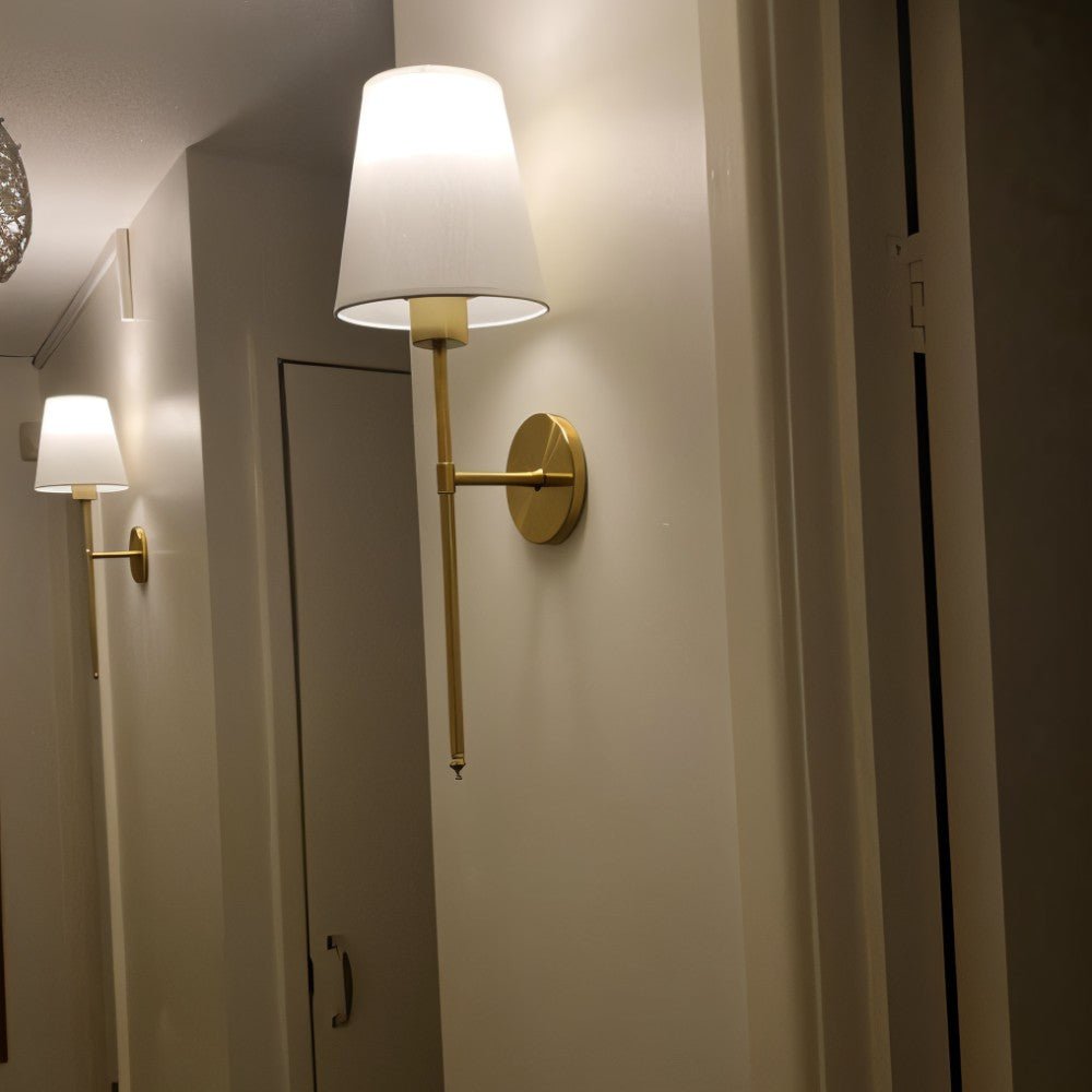 Clérachic brass-gold wall sconce with a white fabric shade lit warm on a narrow hallway wall, with a second one reflected.