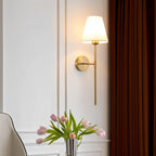 Clérachic gold wall sconce with a white tapered fabric shade on a panelled wall beside a rust curtain and a vase of tulips.