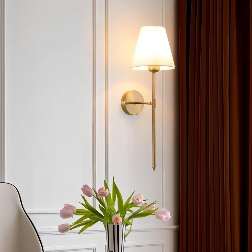 Clérachic gold wall sconce with a white tapered fabric shade on a panelled wall beside a rust curtain and a vase of tulips.