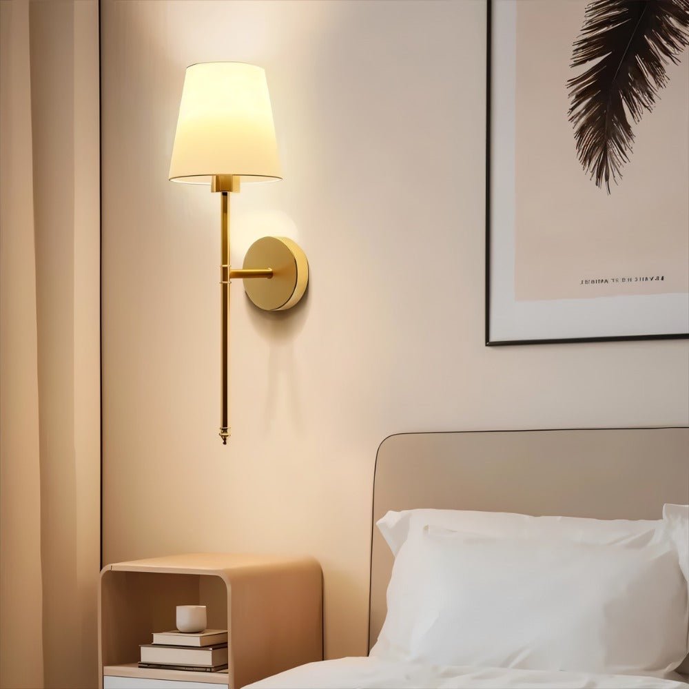 Clérachic gold wall sconce with a white fabric shade glowing over a bedside table and a framed feather print.