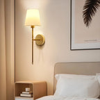 Clérachic gold wall sconce with a white fabric shade glowing over a bedside table and a framed feather print.