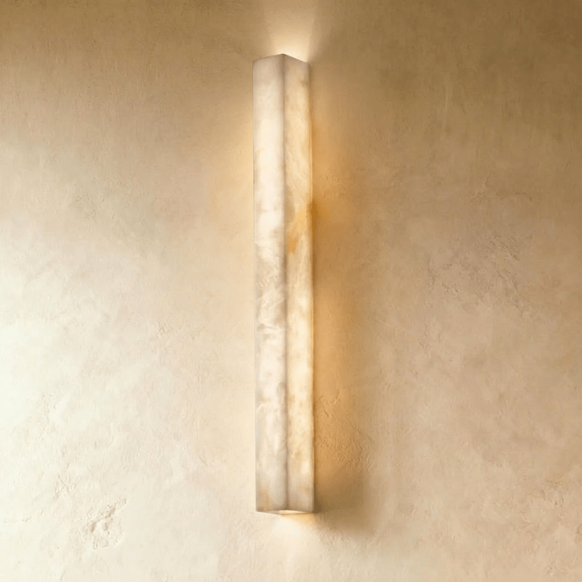 Clérachic longer alabaster bar wall light, lit, its veined stone casting a soft glow above and below on a plaster wall