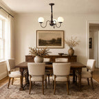 Three white glass globes on curved dark arms, a Clérachic chandelier hung above a long wooden dining table with upholstered chairs.