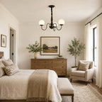 A three-globe Clérachic chandelier hung above a bedroom, with a linen bed, a wooden chest of drawers and a landscape painting on the wall.