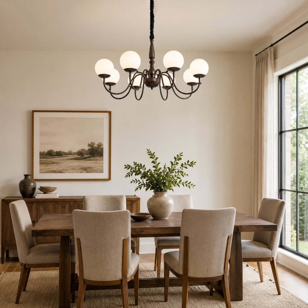 An eight-globe Clérachic chandelier in dark bronze hangs above a wooden dining table with linen chairs and a framed landscape.