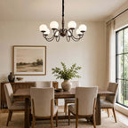An eight-globe Clérachic chandelier in dark bronze hangs above a wooden dining table with linen chairs and a framed landscape.