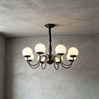 A Clérachic chandelier with eight lit globes hangs in a room with grey plaster walls and ceiling, its dark chain wrapped in black.
