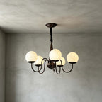A five-globe Clérachic chandelier in dark bronze lit against grey plaster walls, with a black chain and a turned central stem.