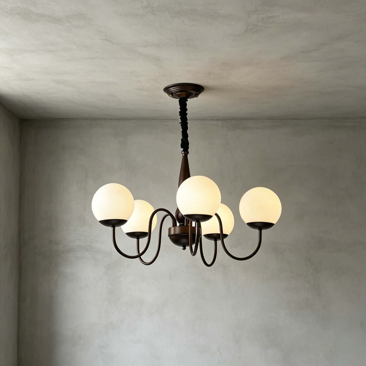 A five-globe Clérachic chandelier in dark bronze lit against grey plaster walls, with a black chain and a turned central stem.