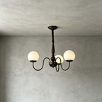 A three-globe Clérachic chandelier with swooping dark arms and a turned stem, lit in a plain room with grey plaster walls.
