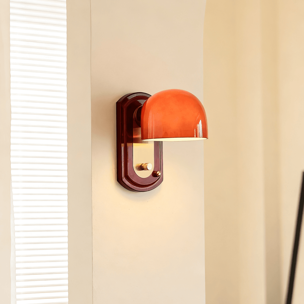 Lit red-orange dome of this Clérachic wall light on a cream wall beside a tall window with white slatted blinds.