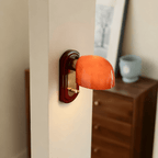 This Clérachic wall light glows orange-red above a walnut chest of drawers, with a framed print and a pale blue pot nearby.