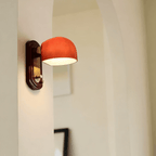 Side-on view of this Clérachic wall light lit in a cream room with an arched opening, its dome glowing orange over a red backplate.