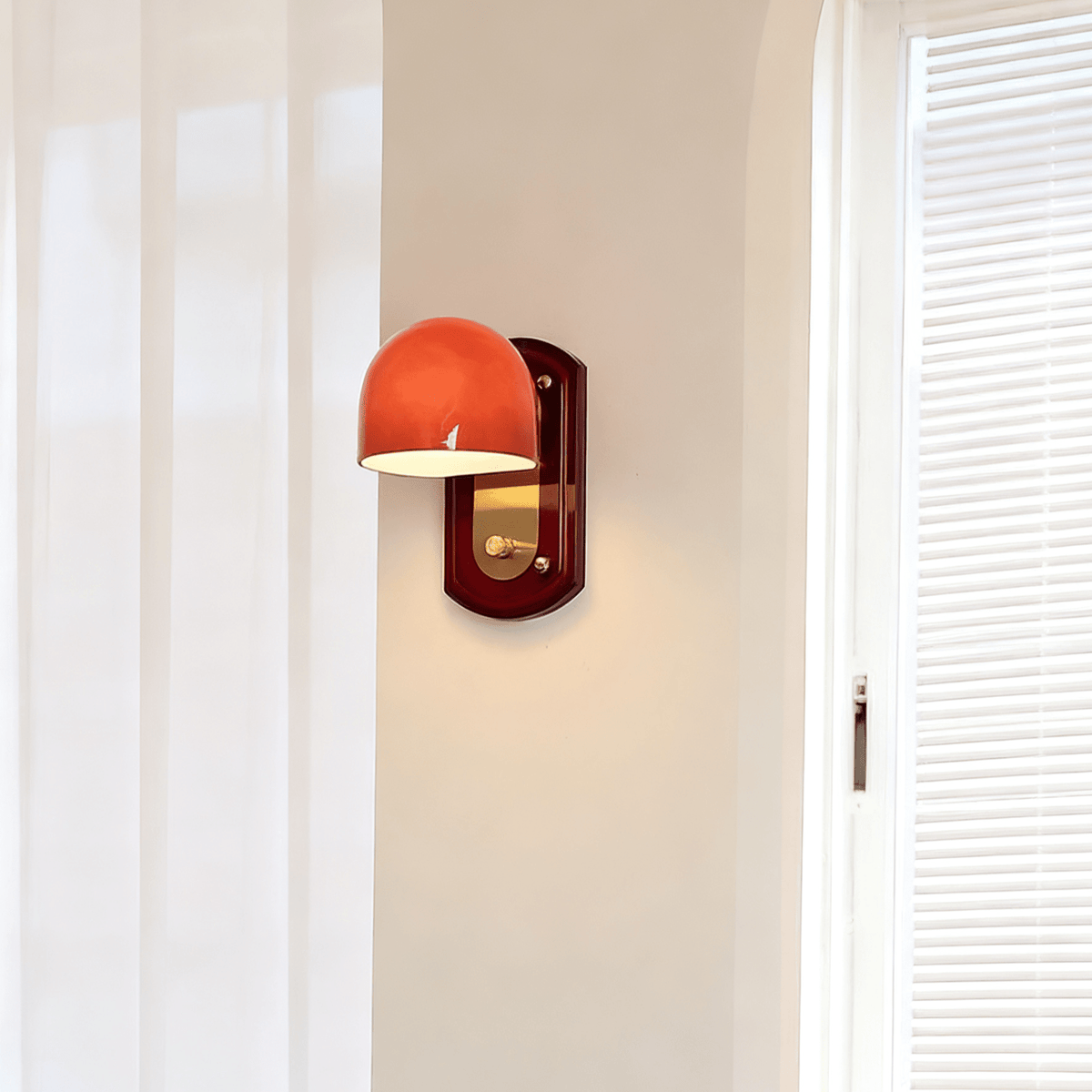 A Clérachic wall light with its dome turned to the left and lit, on a pale wall beside sheer curtains and white blinds.