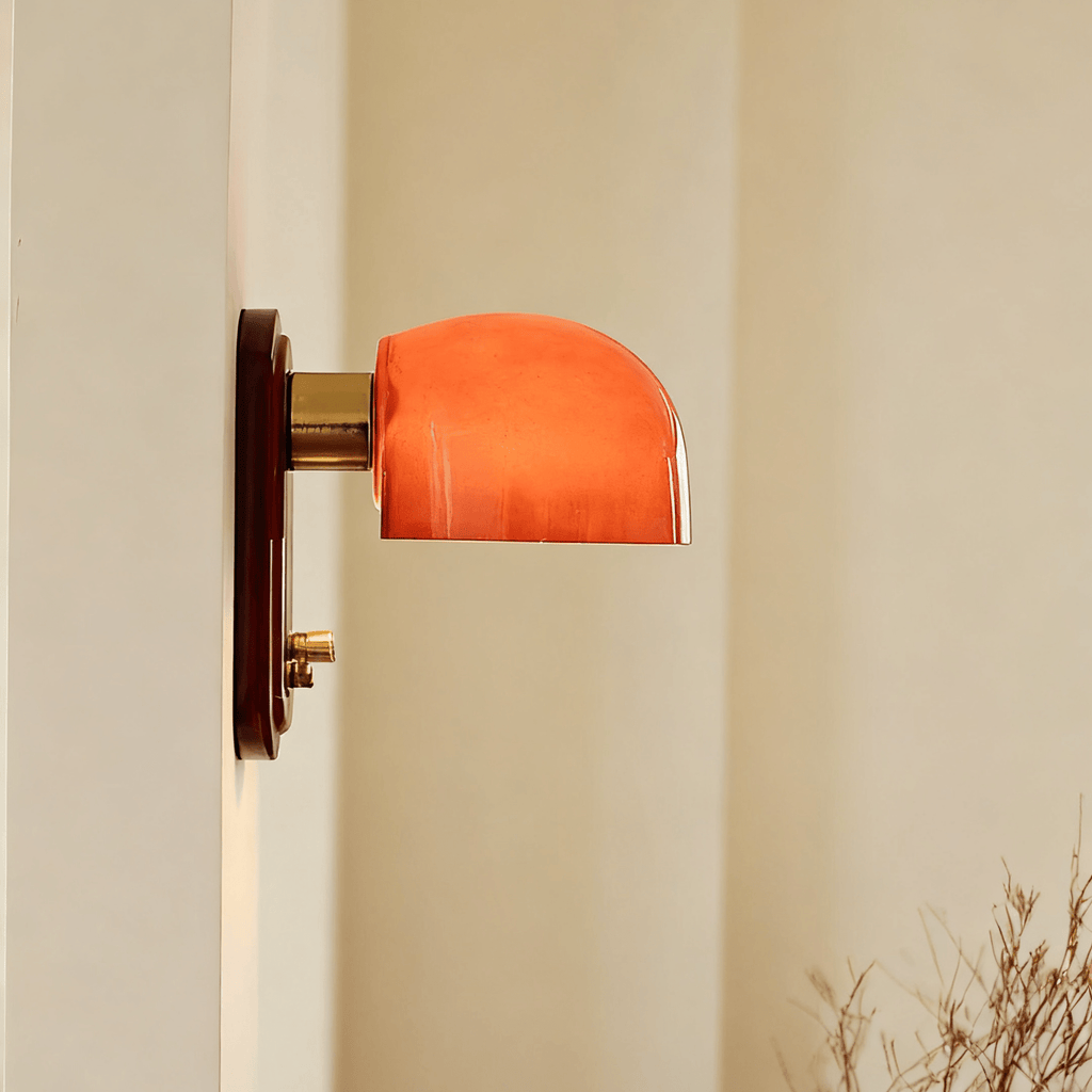 Profile of this Clérachic wall light, its lit orange glass dome on a brass arm projecting from a dark red backplate on a cream wall.
