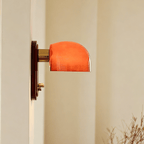 Profile of this Clérachic wall light, its lit orange glass dome on a brass arm projecting from a dark red backplate on a cream wall.