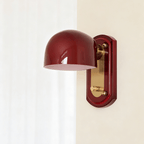 Unlit deep red dome on this Clérachic wall light, with a brass arm and gold-toned panel on its red backplate, in a bright room.