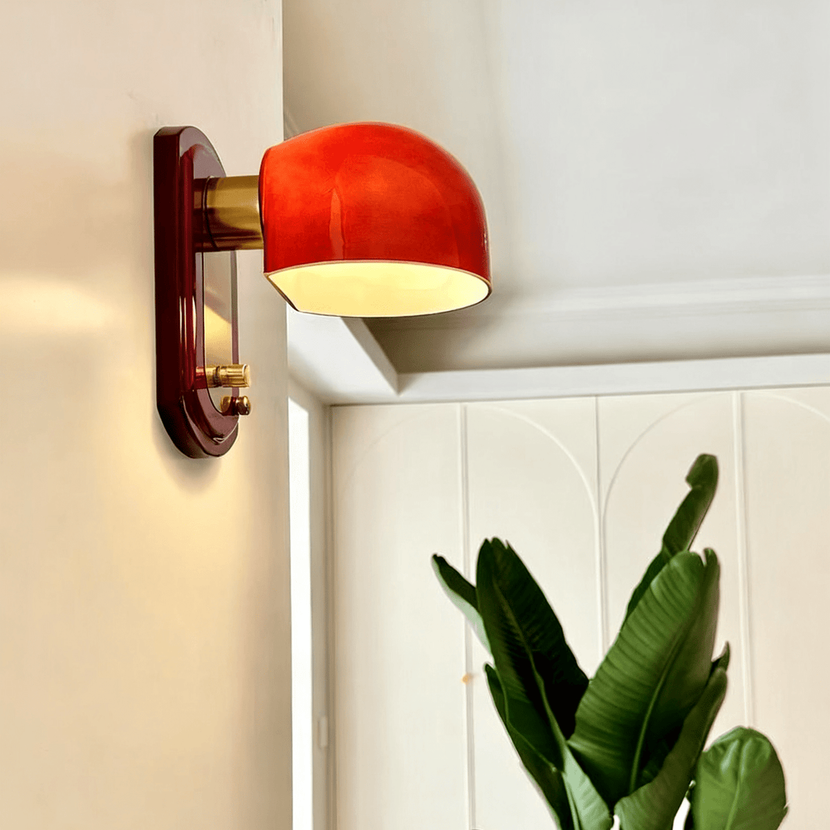 This Clérachic wall light lit in a cream room beside a panelled white door, with the leaves of a green plant in the corner.
