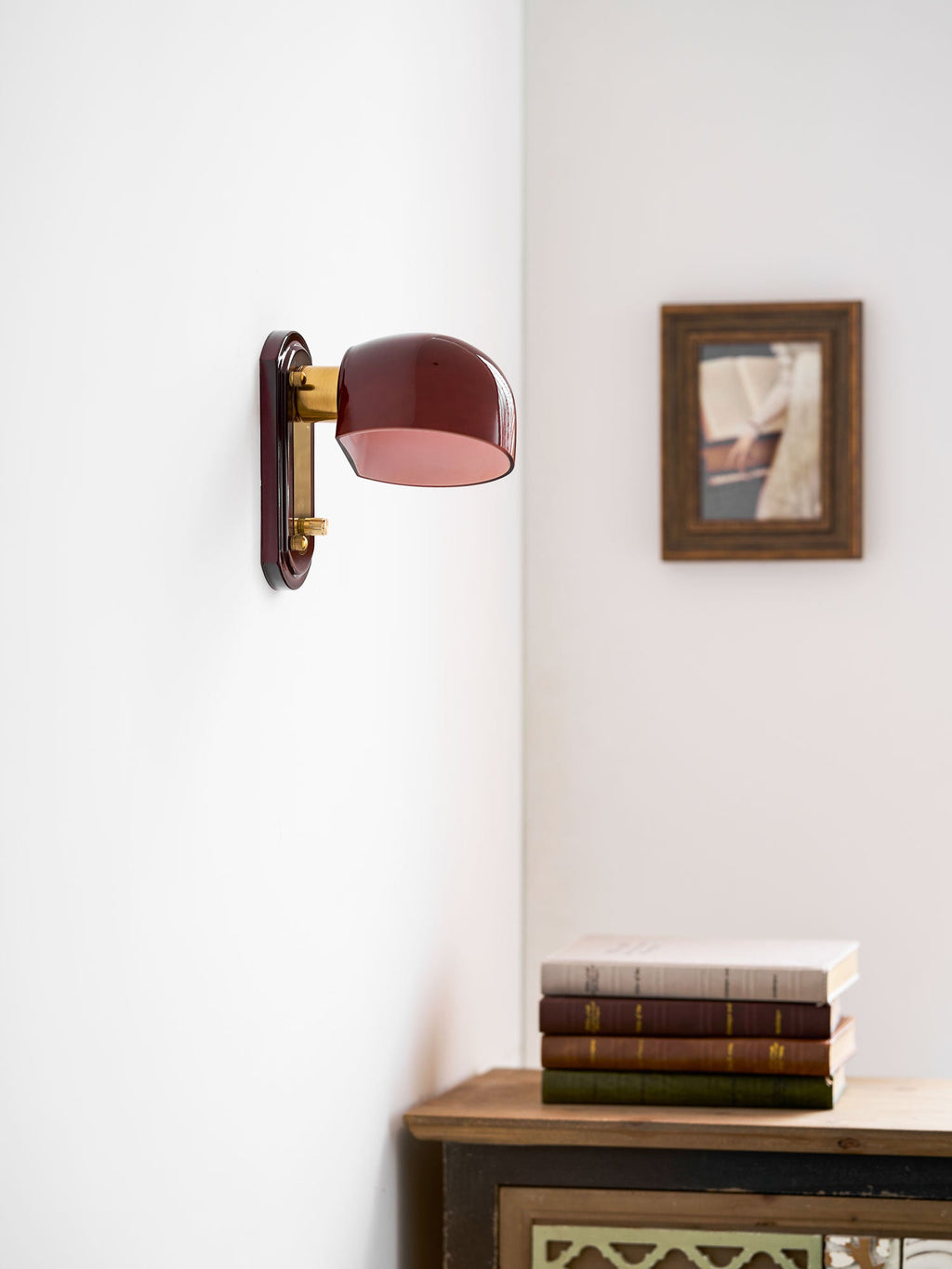 A Clérachic wall light with a dark red dome on a white corner wall, next to a framed picture above a stack of books.