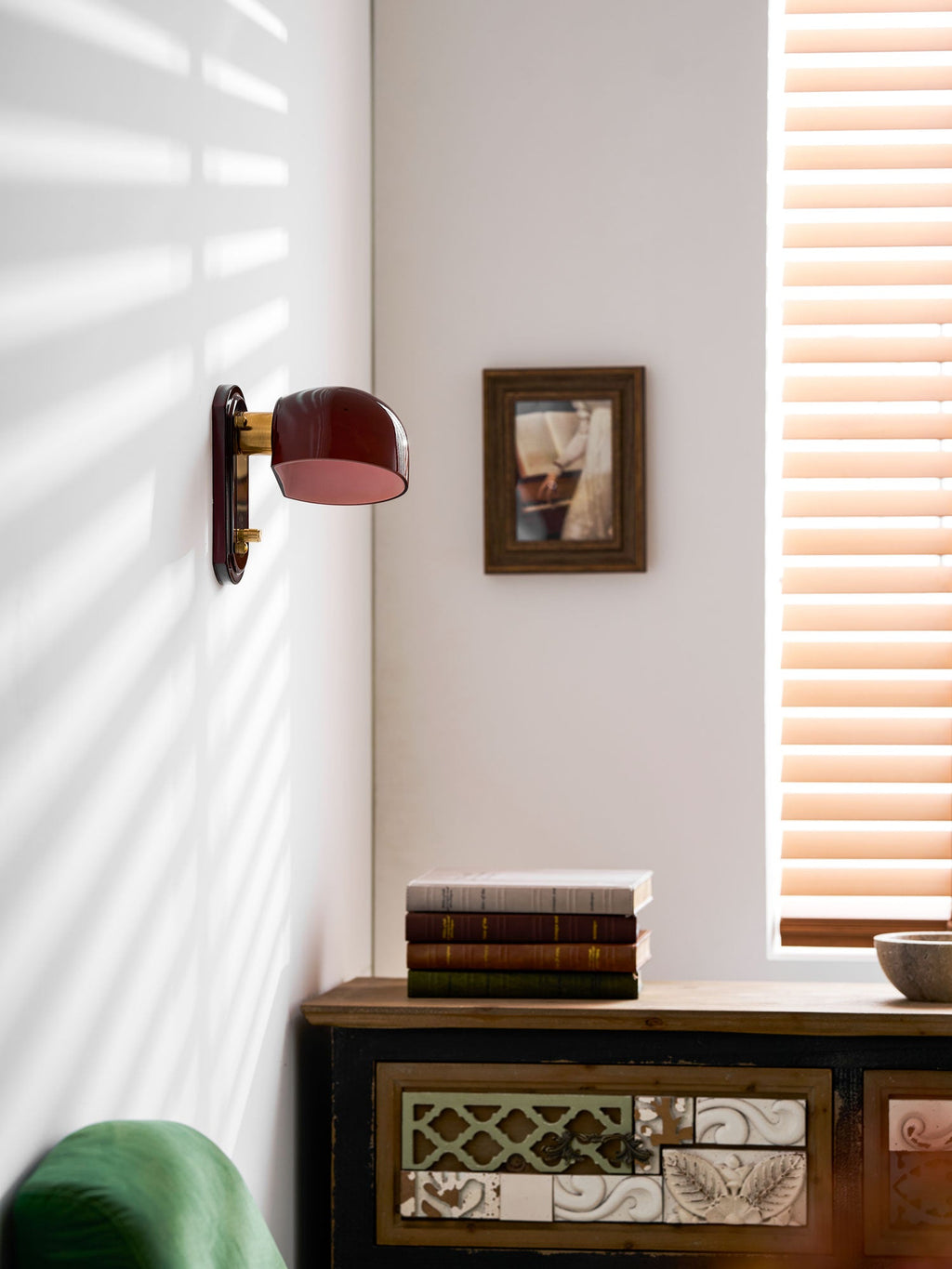 Striped window light falls across a white wall where this Clérachic wall light hangs, above a patterned wooden cabinet and a green chair.