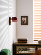 Striped window light falls across a white wall where this Clérachic wall light hangs, above a patterned wooden cabinet and a green chair.