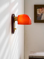 Close side view of this Clérachic wall light lit orange-red, with its brass arm, framed picture and sunlit blind shadows on the wall.