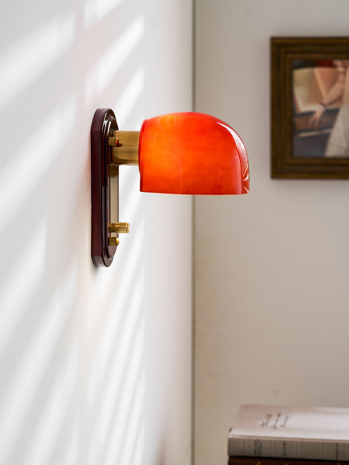 Close side view of this Clérachic wall light lit orange-red, with its brass arm, framed picture and sunlit blind shadows on the wall.