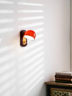 Lit dome of this Clérachic wall light on a white wall striped by blind shadows, above a stack of books on a dark cabinet.