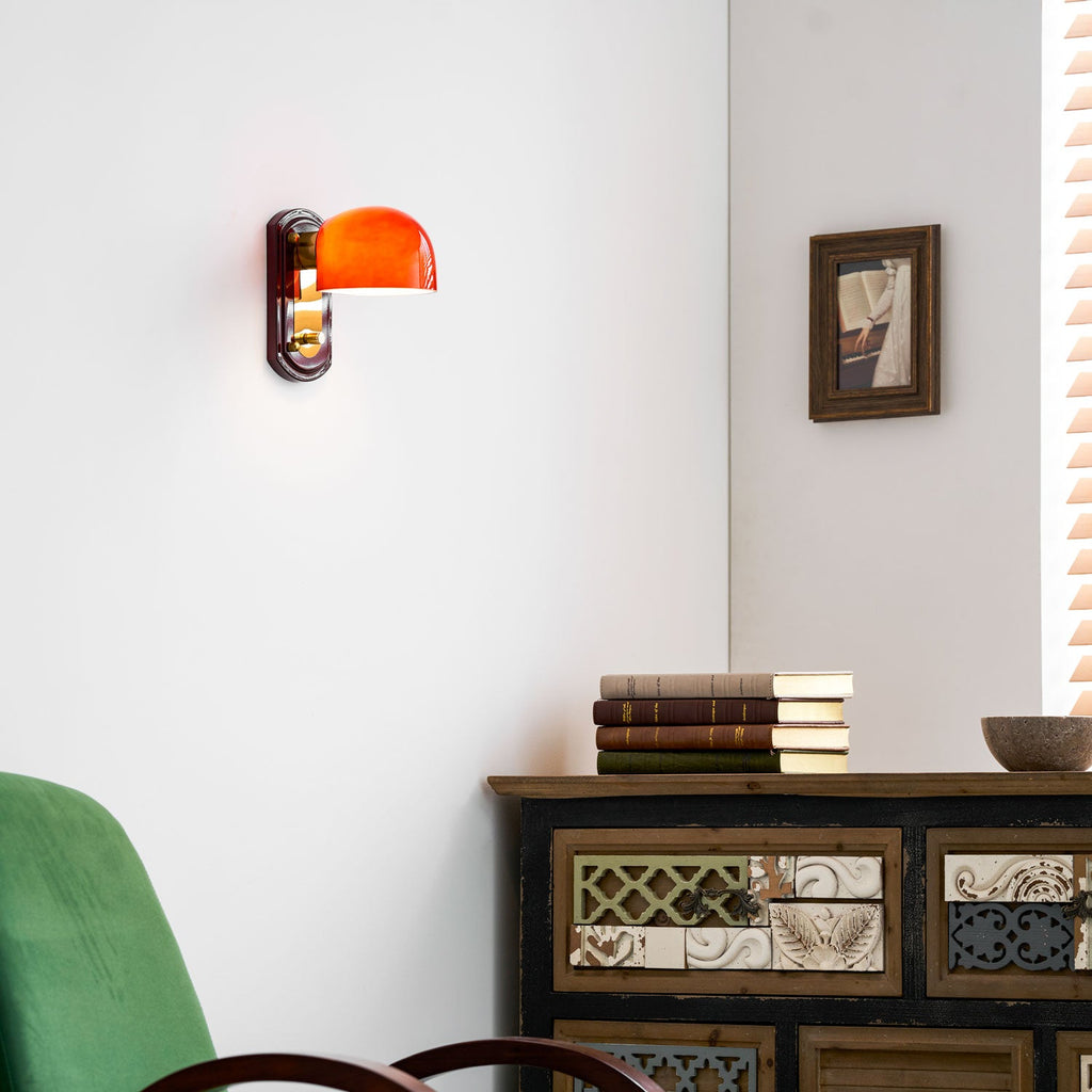 This Clérachic wall light glows orange on a white wall above a carved wooden cabinet, with a green chair and a bowl in view.
