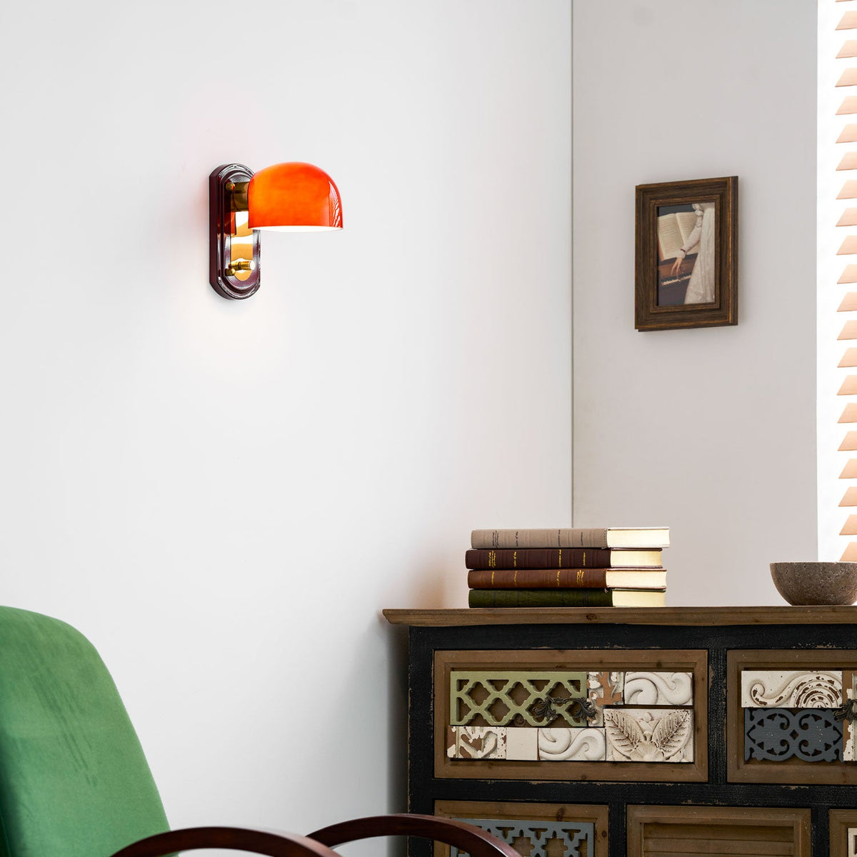 This Clérachic wall light glows orange on a white wall above a carved wooden cabinet, with a green chair and a bowl in view.