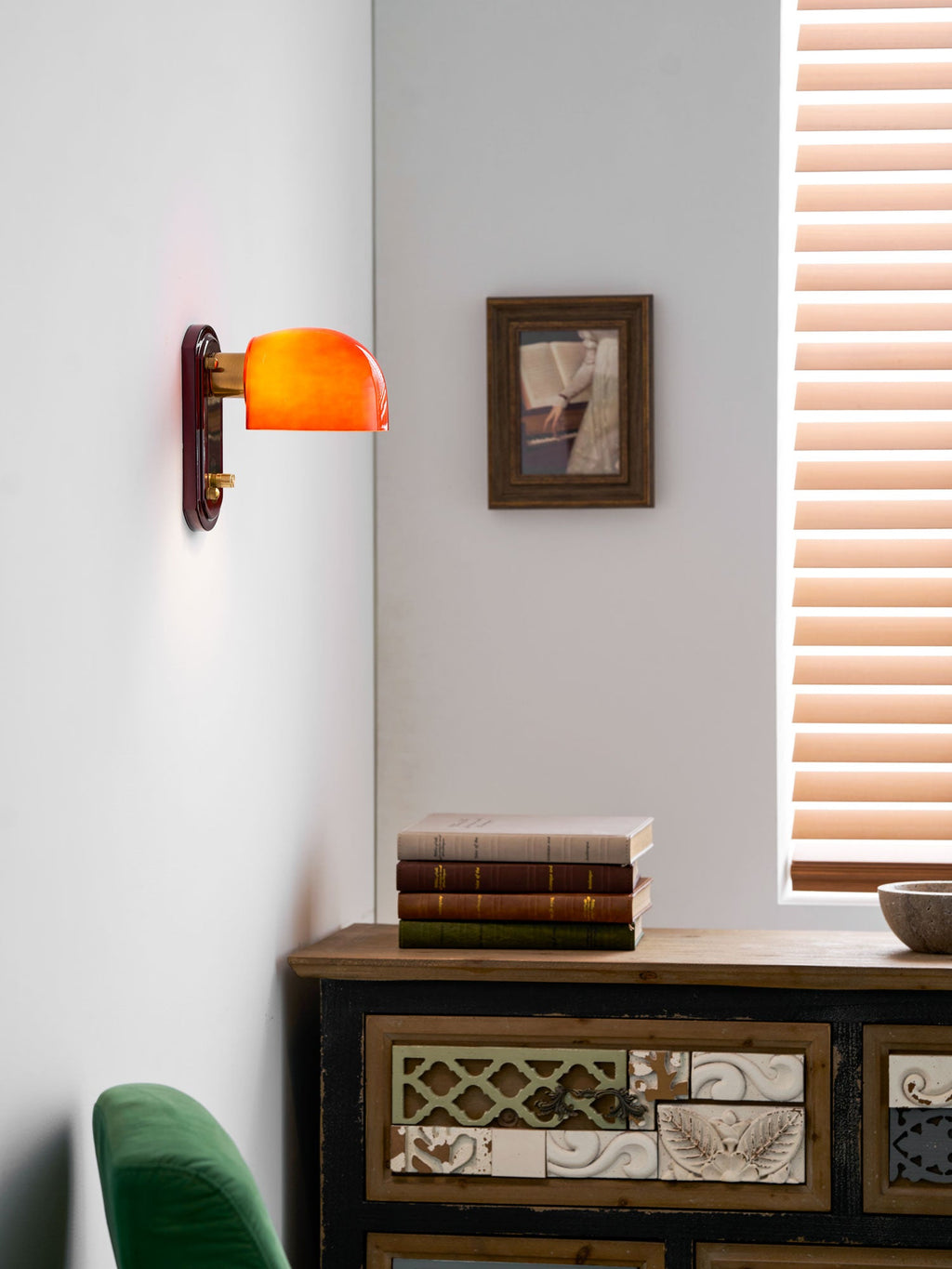 A Clérachic wall light lit amber-orange beside a small framed picture, with a stack of books and a window blind to the right.