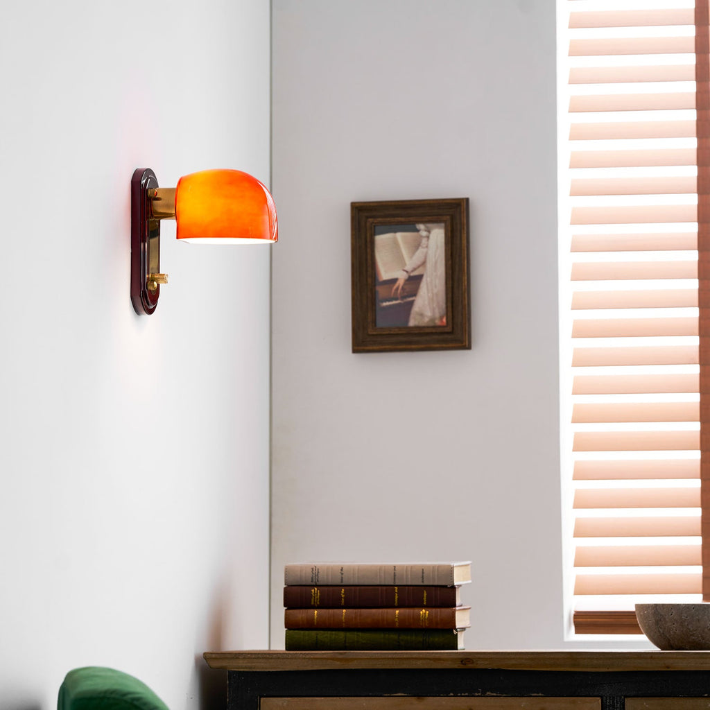 Warm orange glow from this Clérachic wall light on a white wall, with a framed picture, a stack of books and a slatted blind nearby.