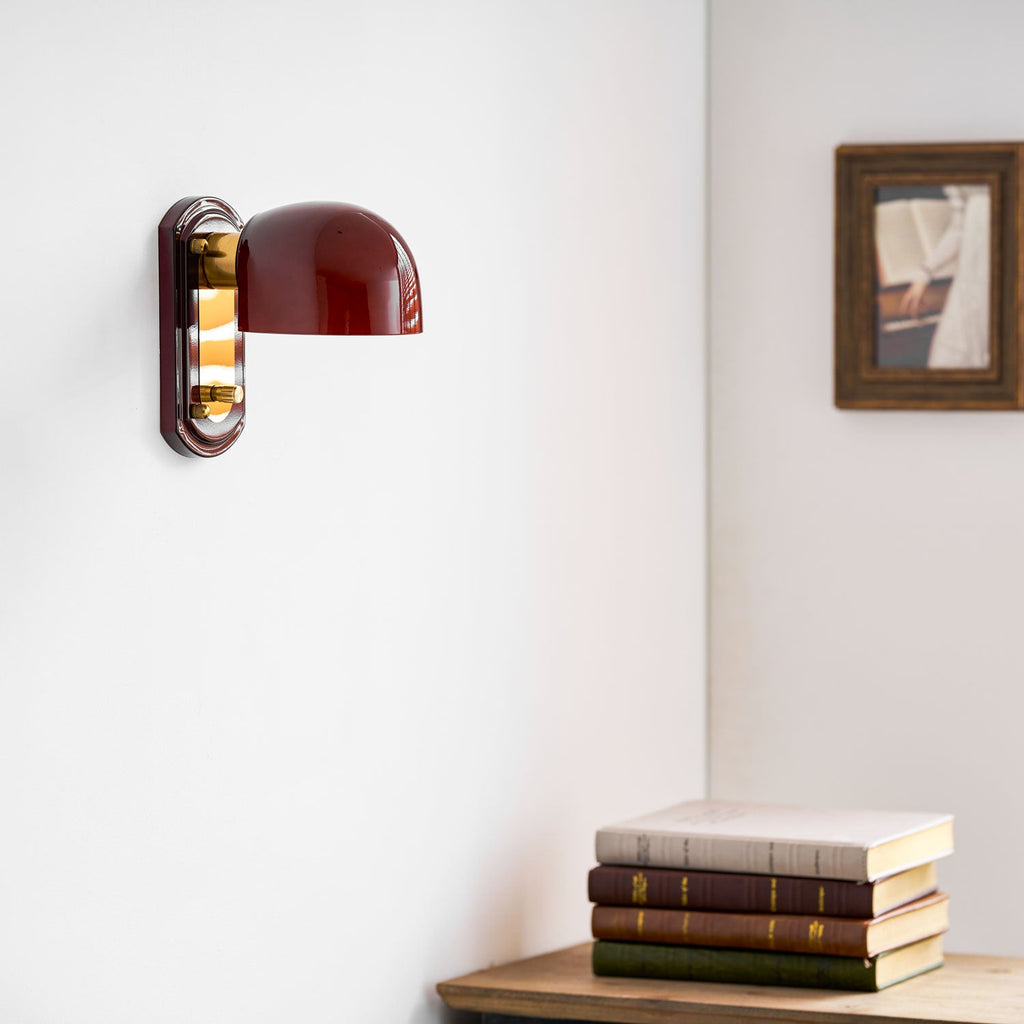 This Clérachic wall light with its dark red dome turned off, beside a framed picture and a stack of books on a wooden shelf.