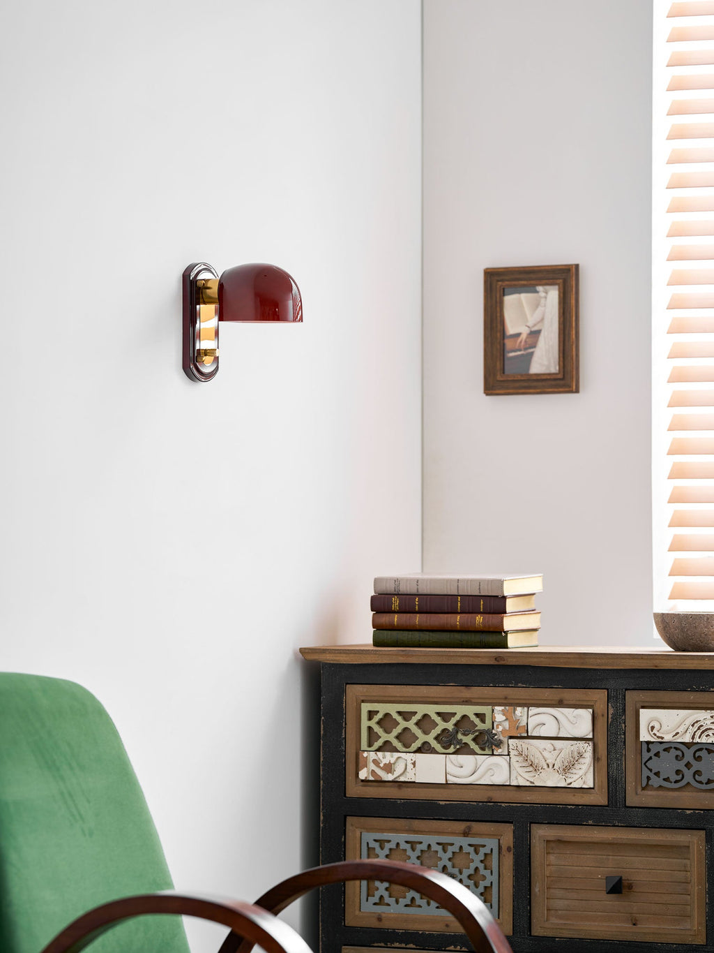 A small, unlit Clérachic wall light seen from further back, above a patterned cabinet with books and a green chair.