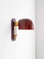 Side view of this Clérachic wall light on a white wall, showing the dark red dome, brass arm and the knob on the red backplate.