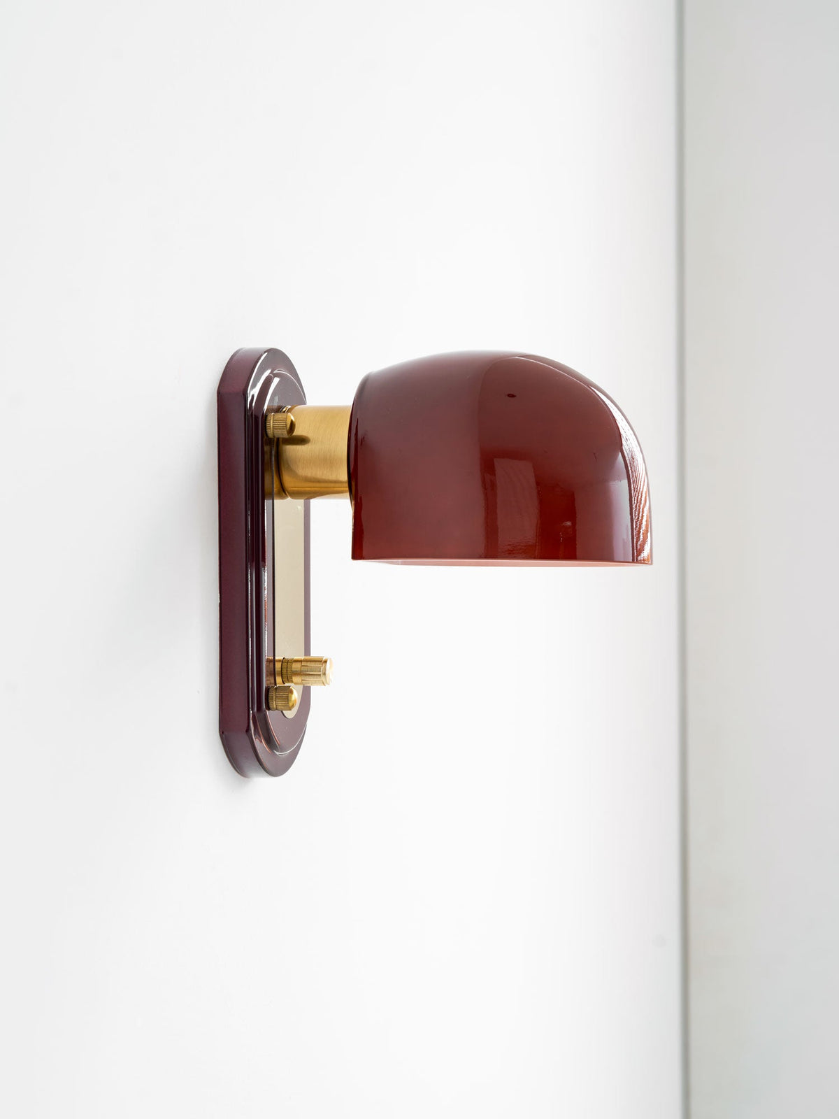 Side view of this Clérachic wall light on a white wall, showing the dark red dome, brass arm and the knob on the red backplate.