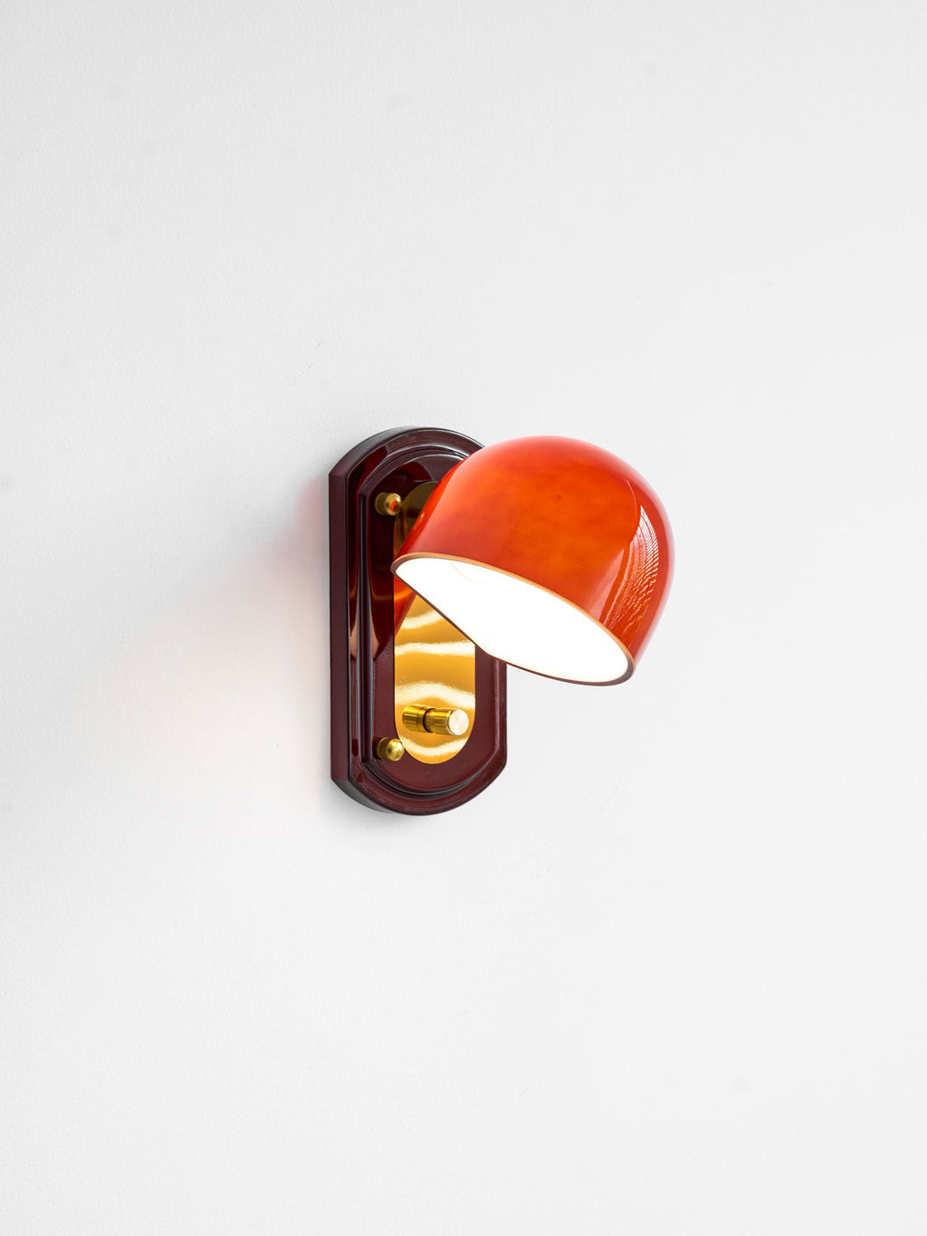 A Clérachic wall light on a white wall with its dome lit and glowing orange-red, turned slightly to the left over a gold panel.