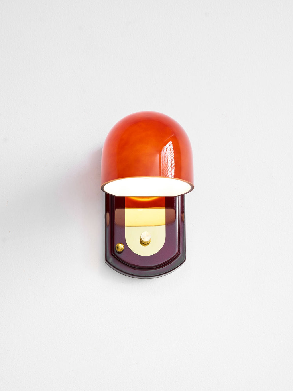 Front view of this Clérachic wall light lit, its glass dome glowing orange-red above the gold panel on the oval backplate.
