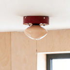 Clérachic red flush mount with a beige glass dome, seen from below against a white ceiling and a birch plywood wall.