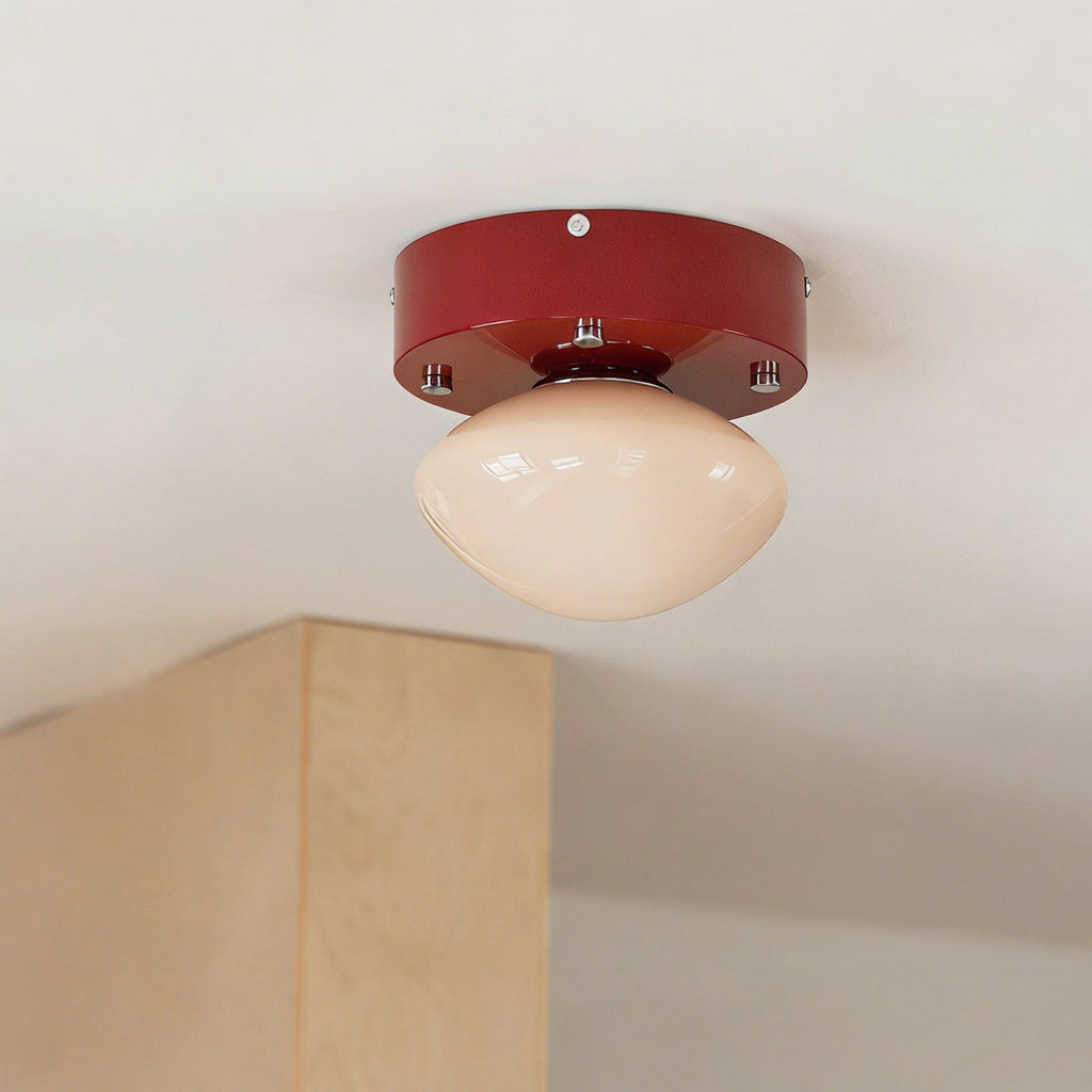 Underside view of this Clérachic flush mount, showing the beige glass dome and the red canopy with its silver bolts, beside a plywood column.