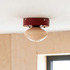 Red-canopied Clérachic flush mount with a beige dome, hanging beside a birch plywood wall and a window.