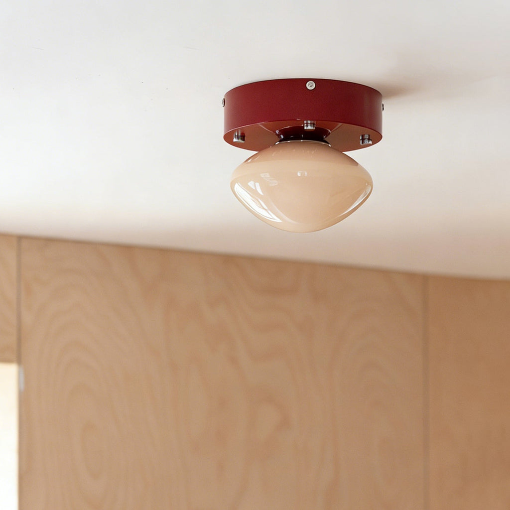 Small red Clérachic flush mount with a beige glass dome, seen from a distance on a white ceiling above a plywood wall.