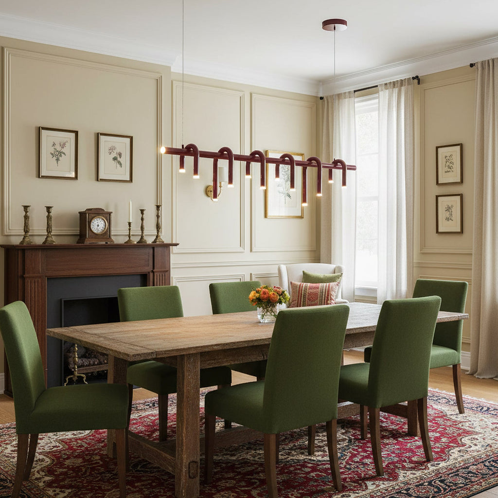 A long red linear Clérachic chandelier above a wooden dining table with green chairs, in a cream panelled room.