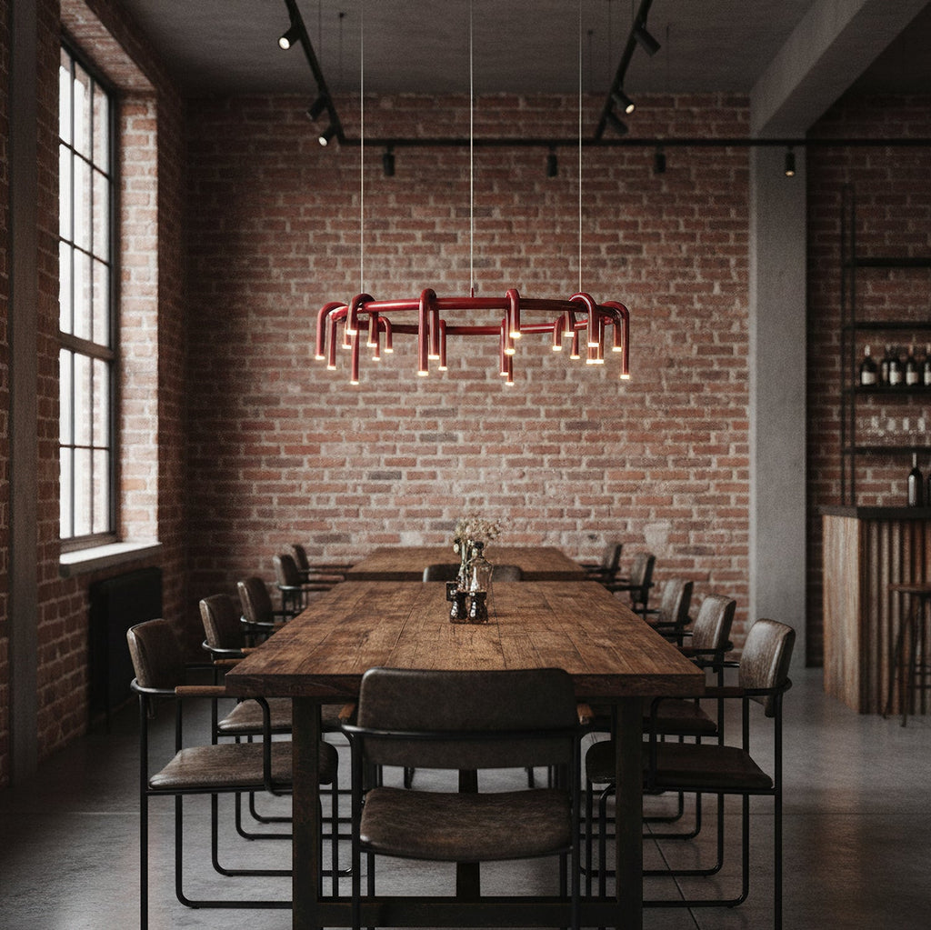 A Clérachic ring chandelier in red over a long wooden table with black-framed chairs, in a brick-walled room with tall windows.