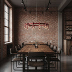 A Clérachic ring chandelier in red over a long wooden table with black-framed chairs, in a brick-walled room with tall windows.