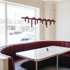 A linear red Clérachic chandelier hangs near a window above a white table and burgundy curved booth seating.